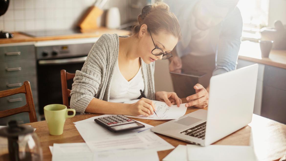 Frau an Tisch mit Laptop, Zetteln, Stift und Taschenrechner