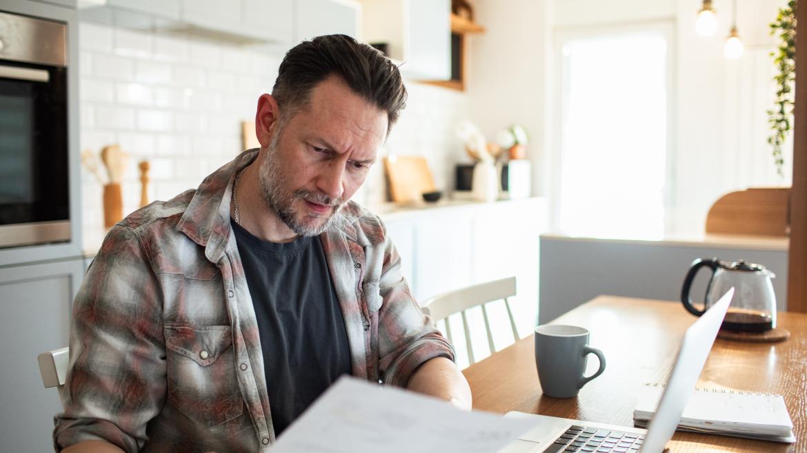 Mann mit Zetteln in Hand sitzt vor Laptop