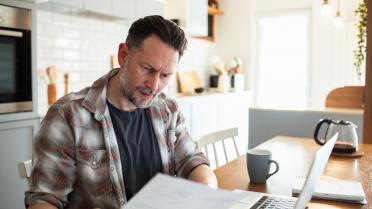 Mann mit Zetteln in Hand sitzt vor Laptop