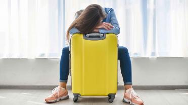 Frau mit gelbem Koffer sitzt am Flughafen. Das Gesicht ist nnicht zu sehen, der Kopf ruht auf dem gelben Koffer.