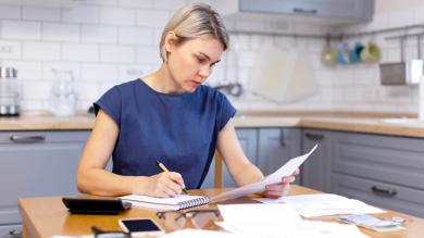 Frau sitzt an Küchentisch mit Papieren, Stift und Zettel