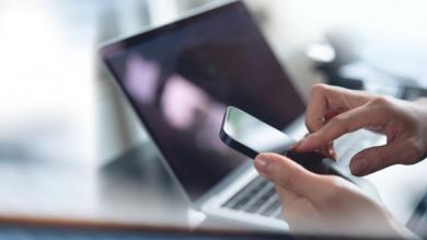 Nahaufnahme Frau tippt auf Smartphone, Laptop im Hintergrund