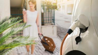 Frau mit Handy in der Hand und weißem Kleid läuft auf ein weisses E-Auto zu, welches an einer Ladesäule geladen wird.