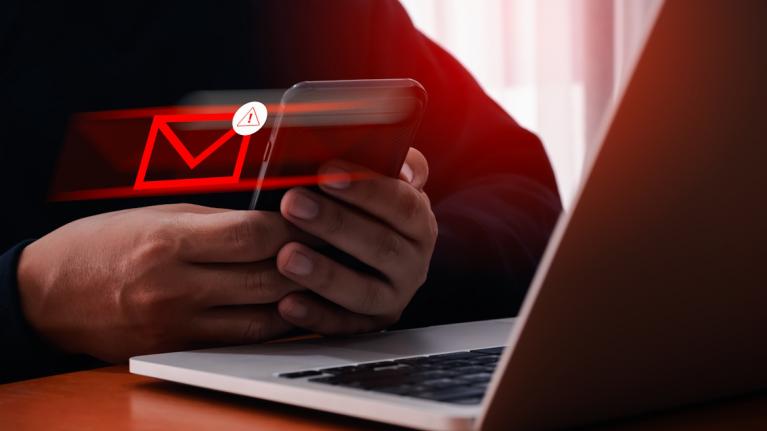 Nahaufnahme Laptop, davor Hände mit Smartphone. Daraus fliegt ein roter Briefumschlag: Phishing Amazon