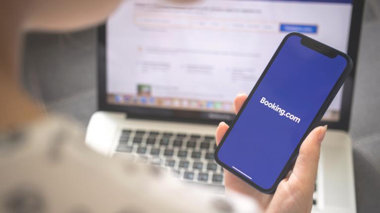Frau mit Smartphone öffnet booking App, im Hintergrund Laptop