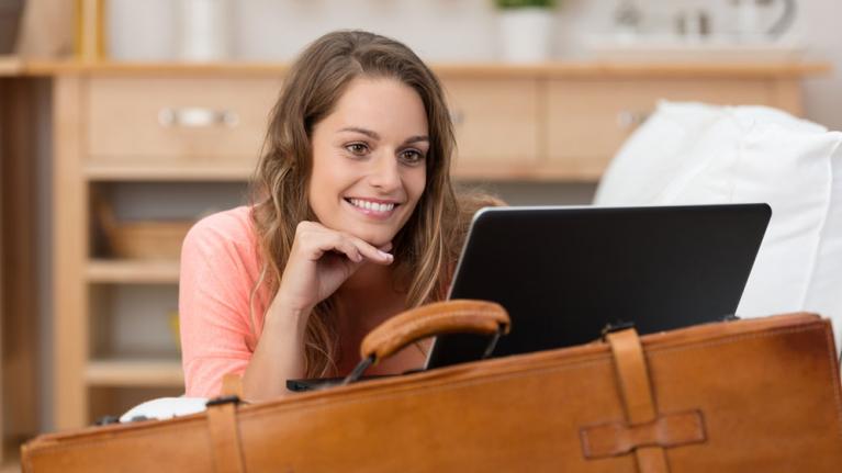 Frau auf Koffer gestützt bucht Urlaub am Laptop