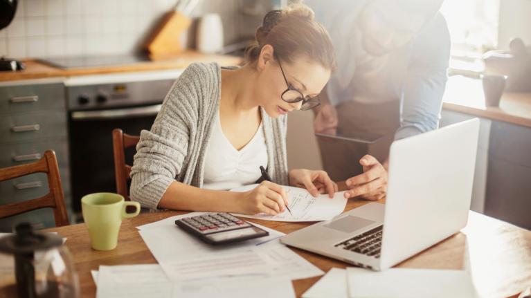 Frau an Tisch mit Laptop, Zetteln, Stift und Taschenrechner