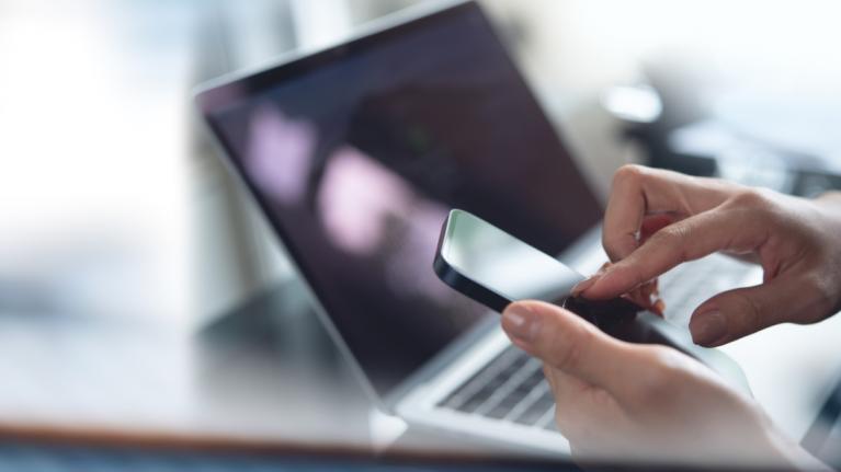 Nahaufnahme Frau tippt auf Smartphone, Laptop im Hintergrund