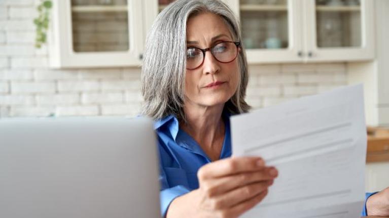 Ältere Frau mit grauen Haaren schaut in der Küche am Laptop auf einen papierrechnung.