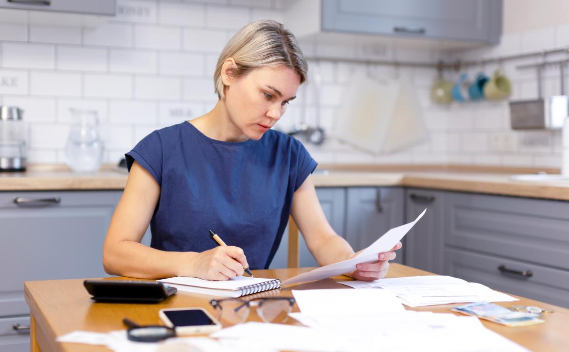 Frau sitzt an Küchentisch mit Papieren, Stift und Zettel