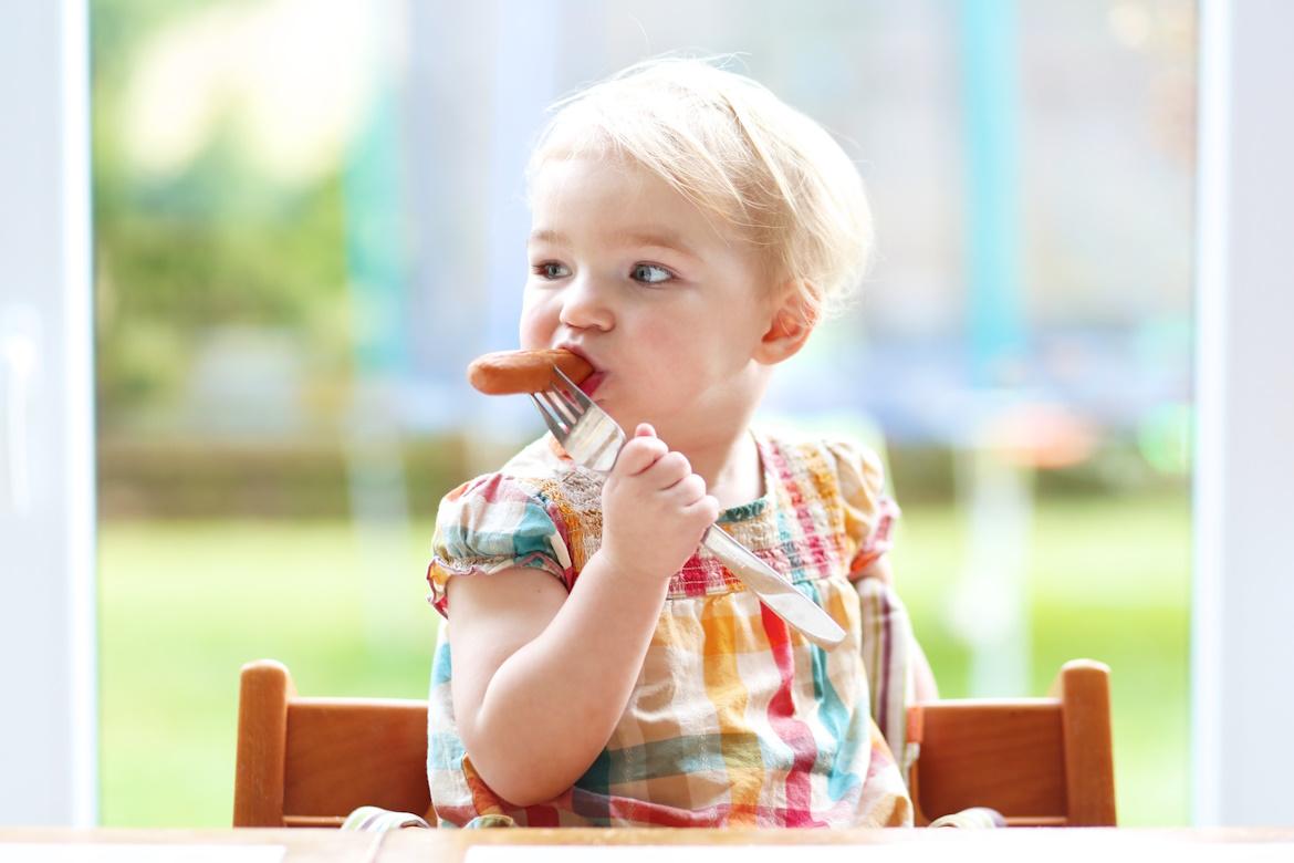 Kleinkind auf Hochstuhl mit Gabel in Hand und Wurst im Mund zu Hygieneschulung Fleisch.