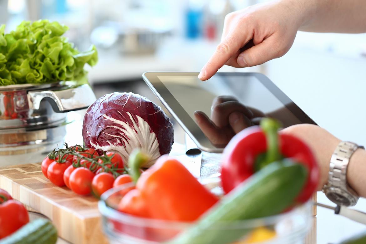 Nahaufnahme Hand zeigt auf Tablet, im Vordergrund verschiedenes Gemüse