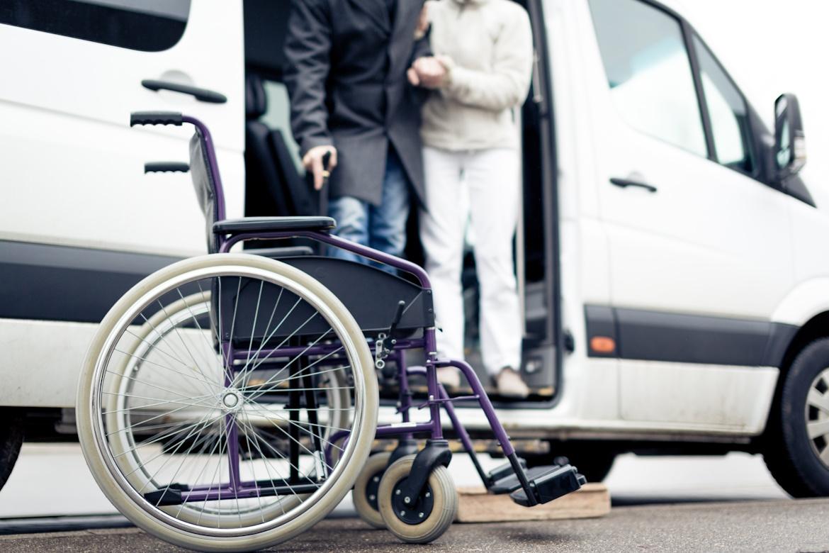 Rollstuhl, im Hintergrund steigt ältere Person mit Hilfe aus einem Bus