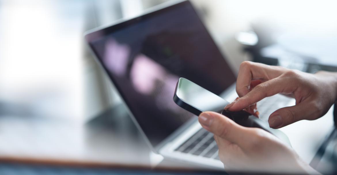 Nahaufnahme Frau tippt auf Smartphone, Laptop im Hintergrund