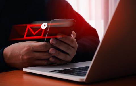 Nahaufnahme Laptop, davor Hände mit Smartphone. Daraus fliegt ein roter Briefumschlag: Phishing Amazon