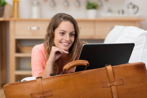 Frau auf Koffer gestützt bucht Urlaub am Laptop