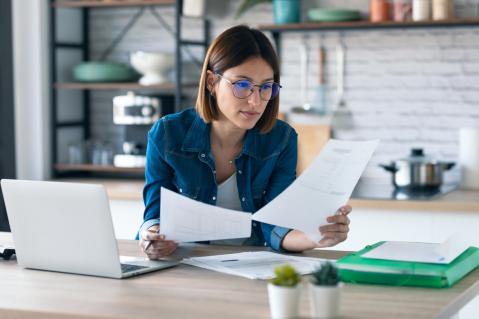 Frau vor dem Computer schaut auf Zettel zu Artikel Kostenfreie Selbstauskunft - Dienstleister lassen sich Service bezahlen