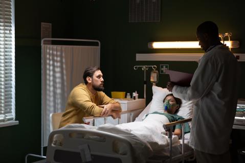 Frau liegt im Krankenbett, Partner an ihrer Seite, rechts ist ein Arzt zu sehen