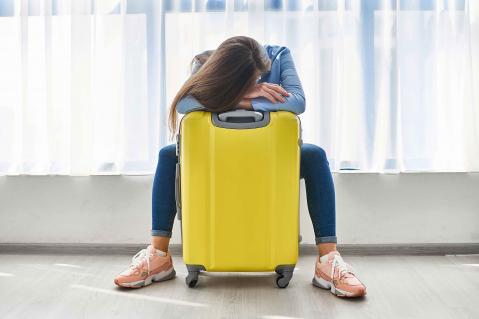 Frau mit gelbem Koffer sitzt am Flughafen. Das Gesicht ist nnicht zu sehen, der Kopf ruht auf dem gelben Koffer.